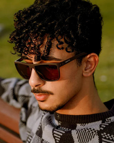 A young man with curly hair wearing stylish sunglasses, sitting outdoors with a thoughtful expression.