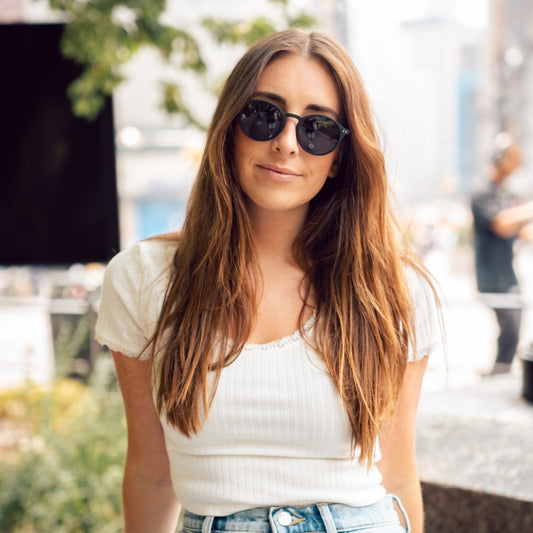 A woman with long hair wearing sunglasses and a white top, standing outdoors.